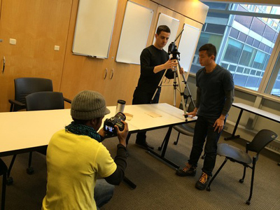 A cameraman taking a photo of two students setting up a camera and tripod.