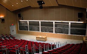 Tiered classroom with red tablet armchairs. Five sliding blackboards and a table are at the front of the classroom.