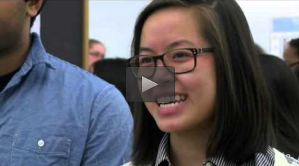 A young woman wearing glasses smiles during an interview while standing in a crowded room.