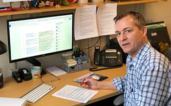 Man sitting at desk, holding a pen, and looking toward camera. Computer and keyboard on desk.