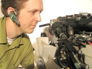 A man wearing a bluetooth earset is crouched next to an internal combustion engine.