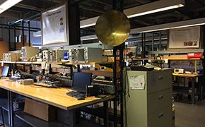 Wooden lab benches with shelves; computer monitors and physics equipment on the on the benches; physics equipment also on the shelves.