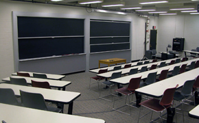 Classroom with rectangular tables and chairs arranged in tiered rows. A small brown table is positioned at the front of the room. It sits in front of two chalkboards.