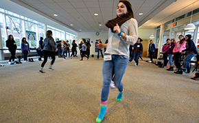 Girls standing in a circle in a large room with tables pushed to the walls. Girls are not wearing shoes, and several girls are running through the center of the circle in an icebreaker activity.