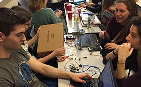 Classroom with students sitting at rectangular tables arranged in rows. Students are working on laptops. Wires and other hardware are on the tables.